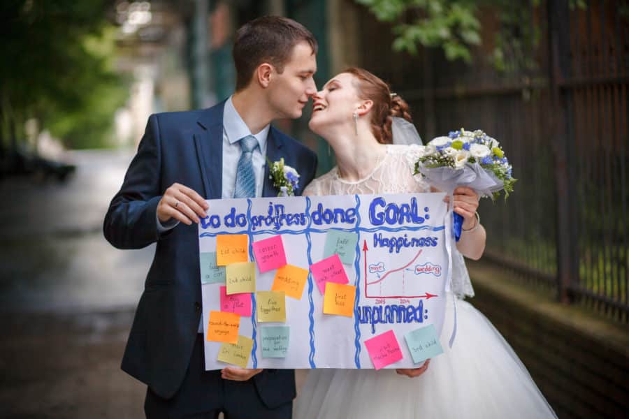 A bride and groom kiss while holding their wedding to do list.