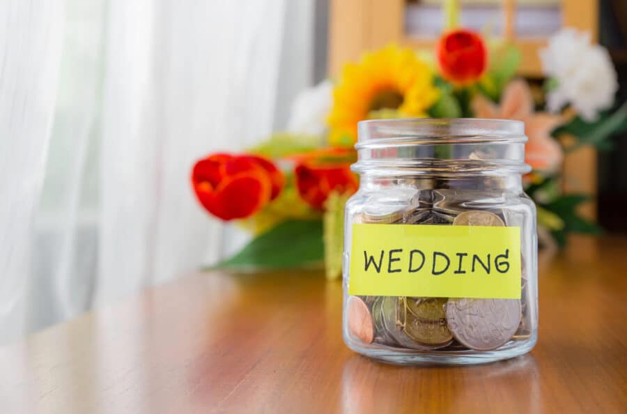 A glass jar with coins on a table. The jar is labeled ‘wedding’.