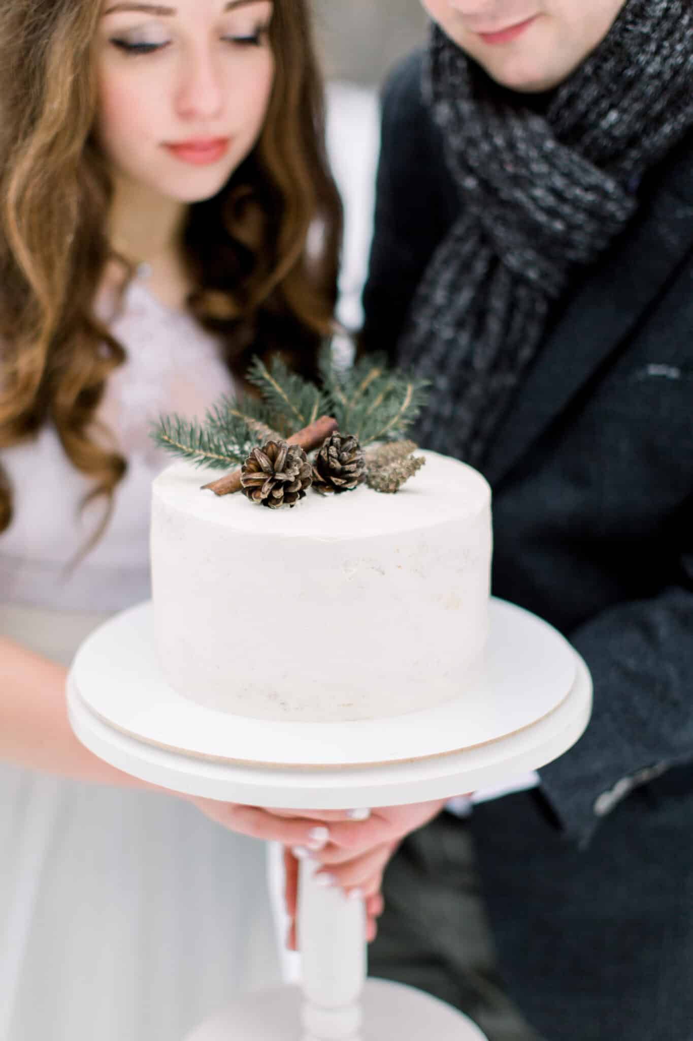 couple holding wedding cake topped with pinecone and winter greenery