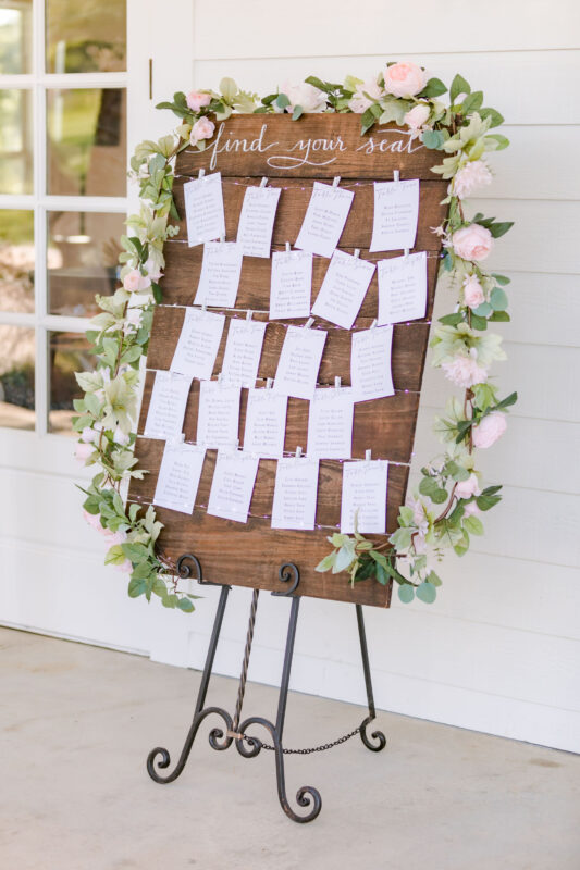 A wooden sign with a simple floral wreath lists the seating assignments for dinner.