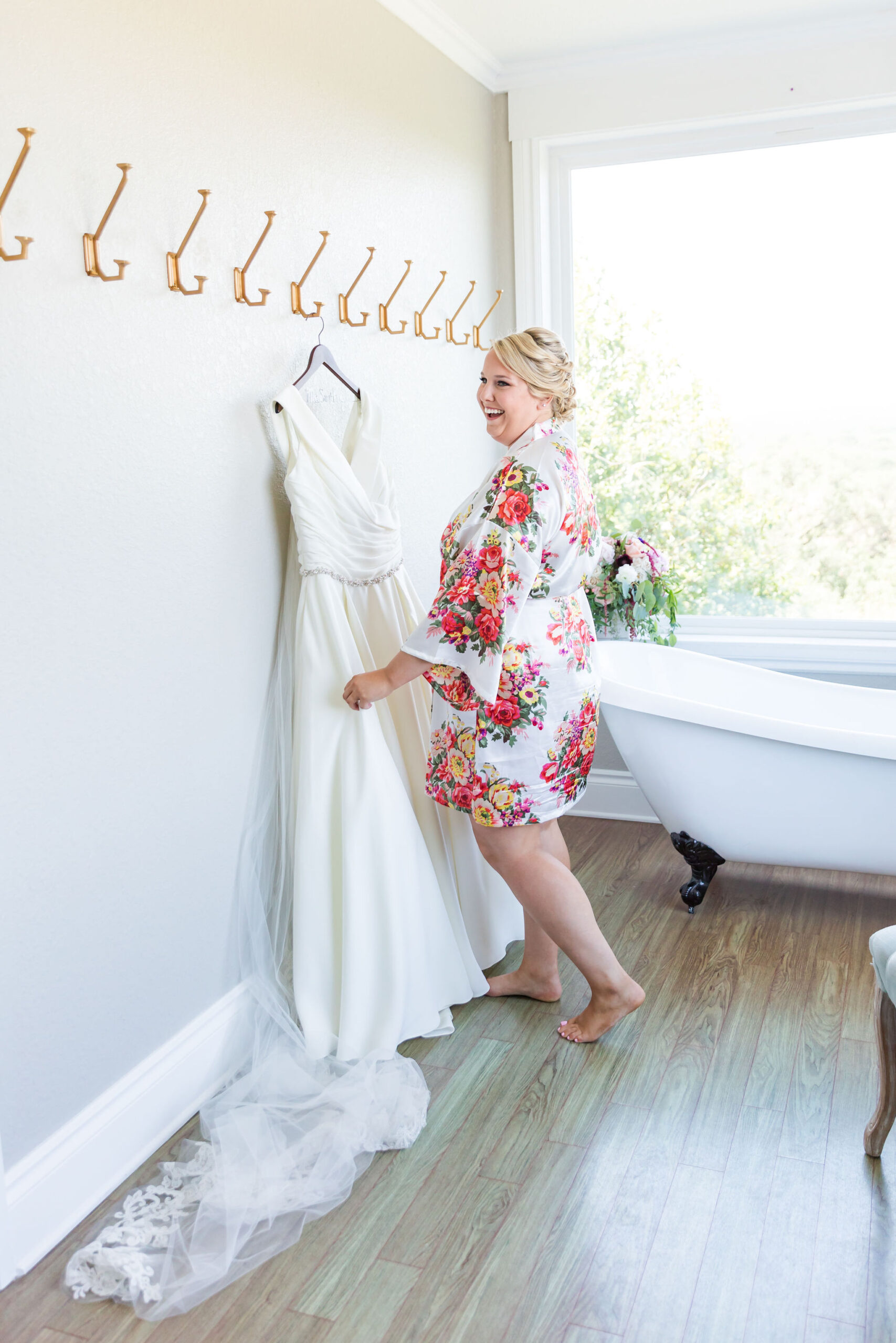 bride in floral robe looking at wedding dress hanging on wall