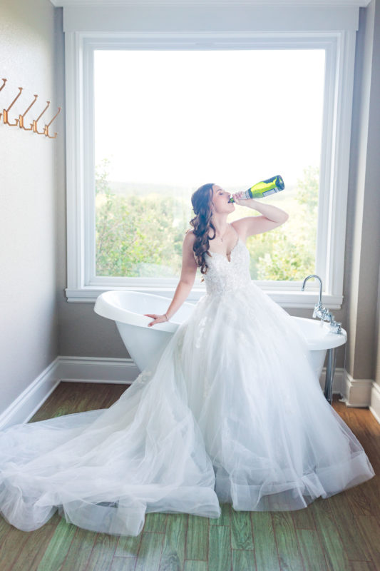 The bride drinks from a champagne bottle by the clawfoot tub in the bridal suite.
