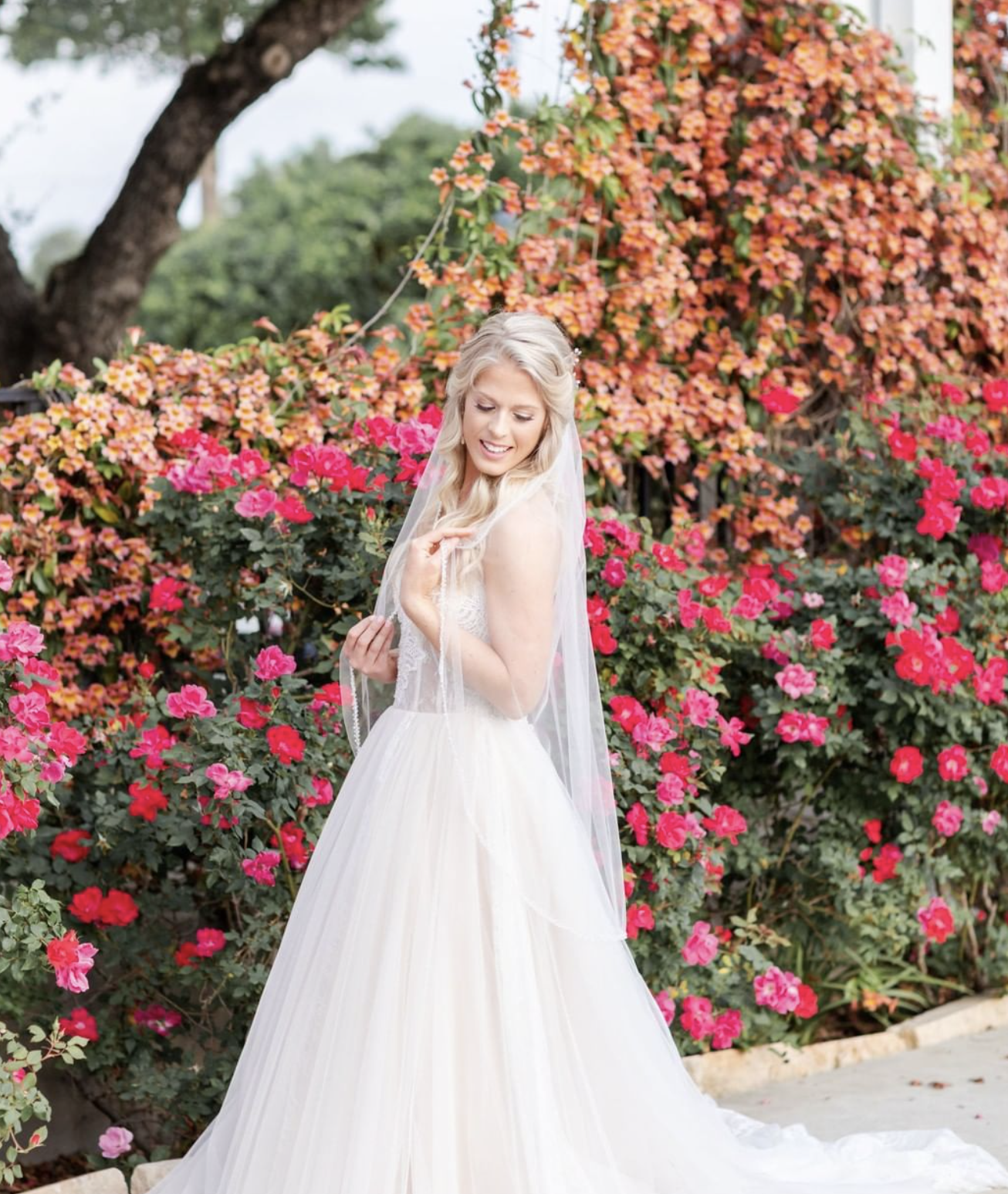 A bride stands in front of a wall of red, orange, and pink flowers with lush green bushes in the background.