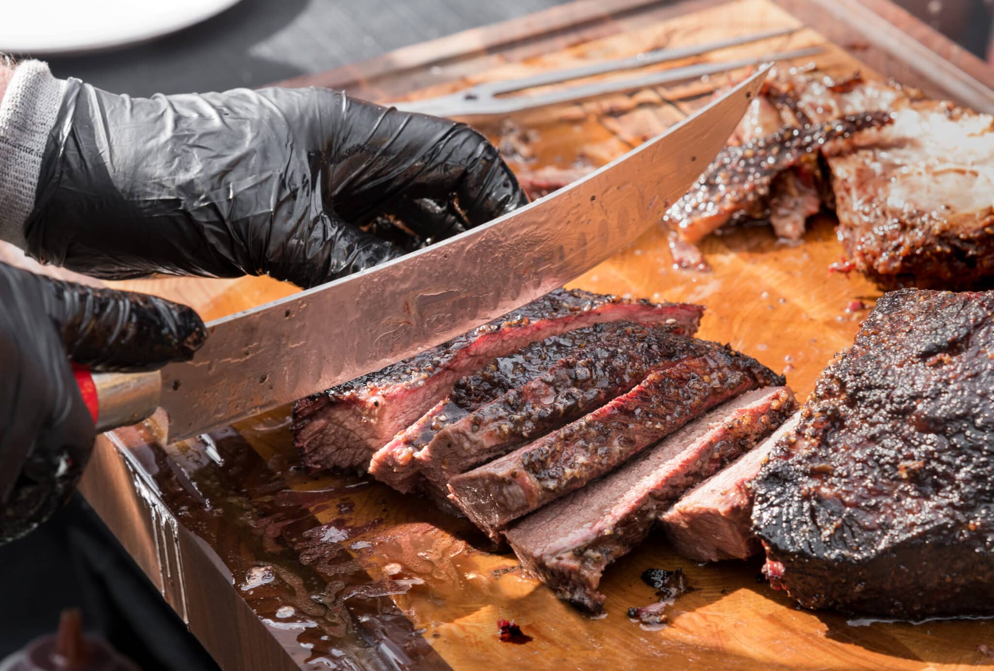 Gloved hands of a chef slicing a portion of delicious juicy tender roast beef brisket with a large knife in a close up on the blade and cutting board